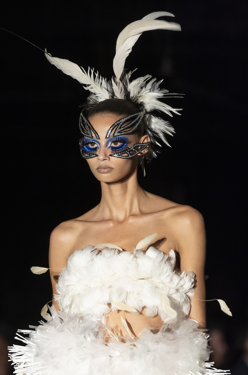 Model wearing a dramatic feathered headdress and a black and blue face mask with feather details, posed with white feathers and fabric, against a dark background.