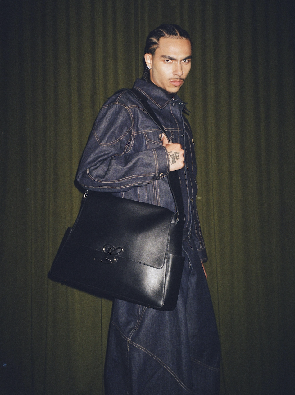 A young man with braided hair, wearing a denim jacket and a black shirt, carrying a black leather bag over his shoulder, standing against a dark curtain background.