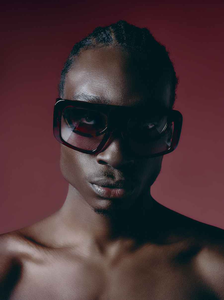 A woman with dark skin wearing large black sunglasses and no shirt, against a red background.