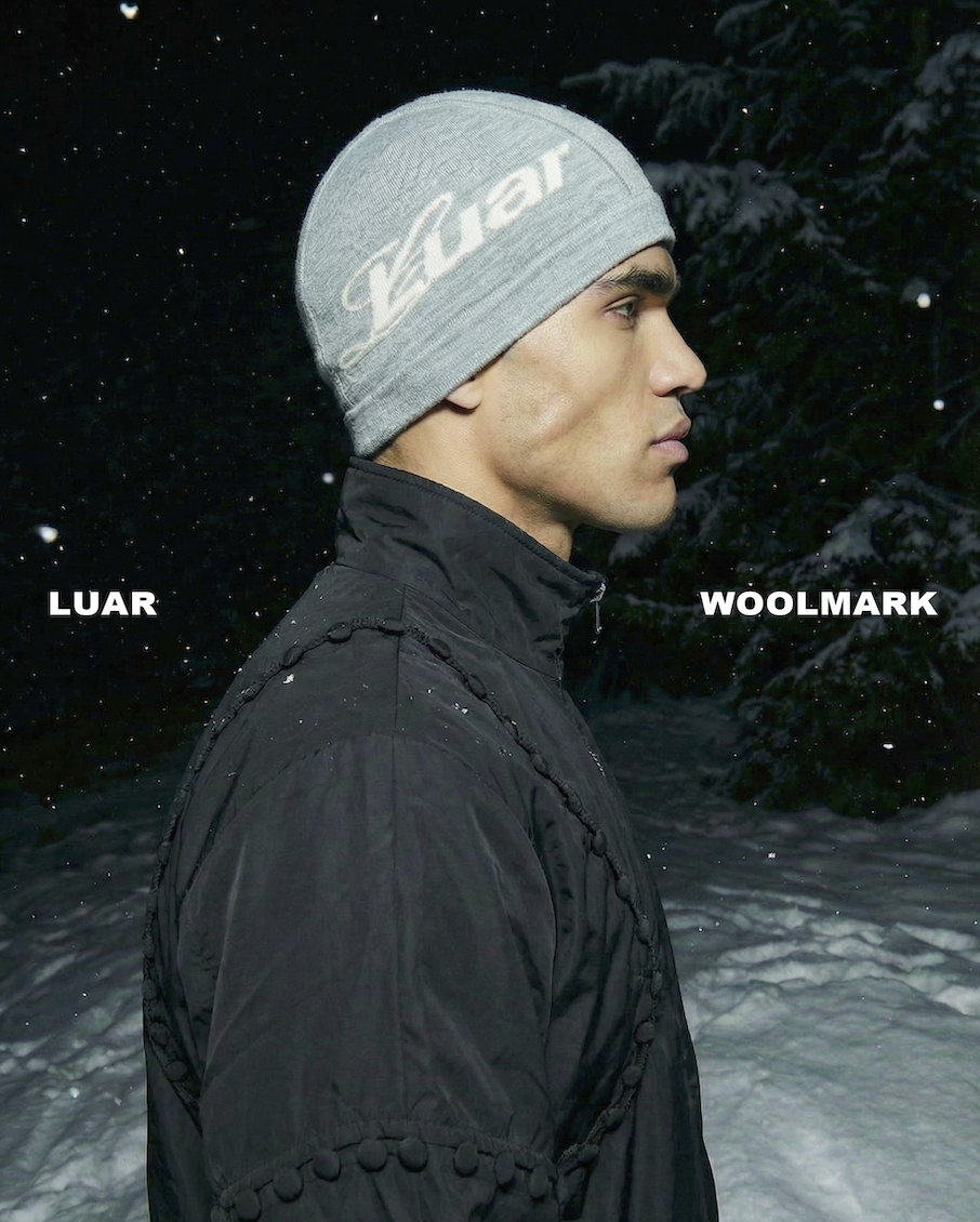 A man in profile view wearing a gray Luar beanie and a black jacket standing in a snowy outdoor area at night with snow-covered trees in the background.