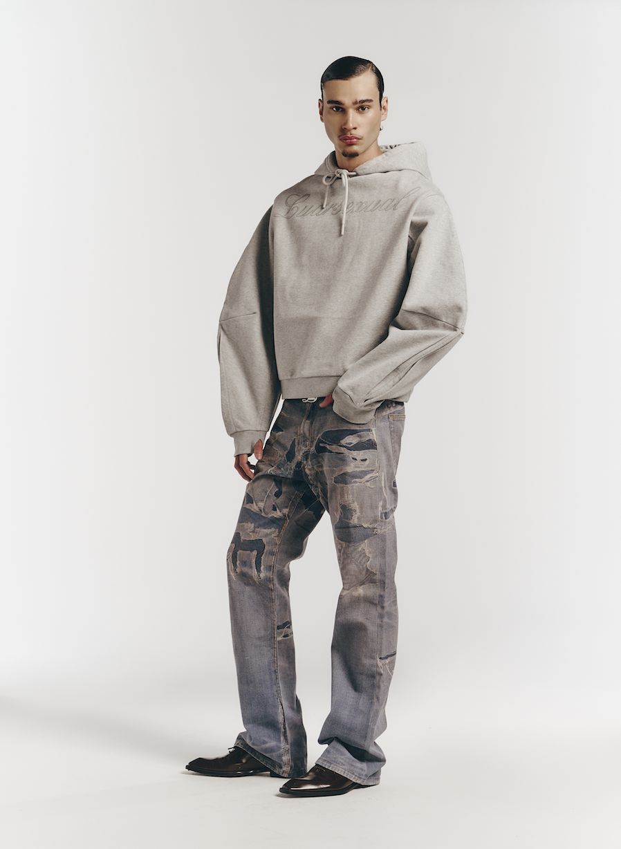 A young man with black hair and a goatee stands in a studio with a white background. He is wearing a gray hoodie, camouflage cargo jeans, and black shoes.