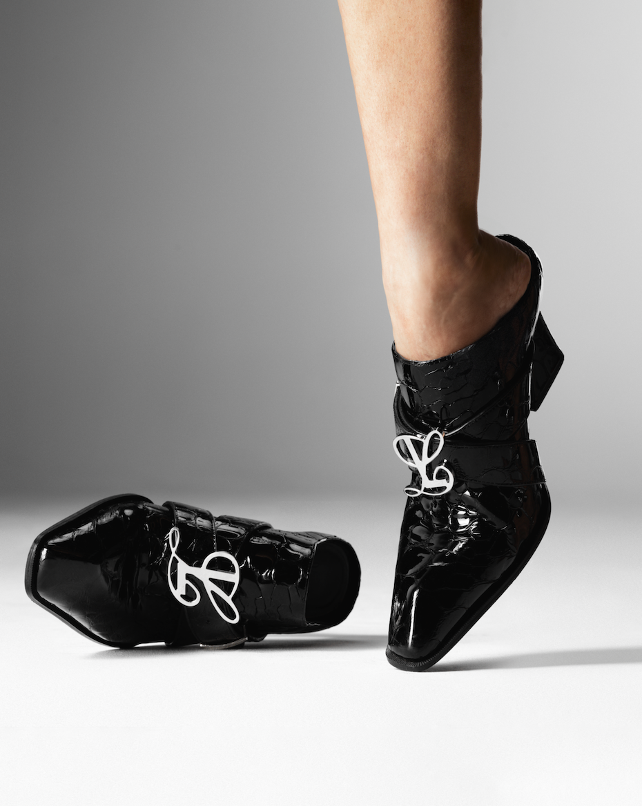 Close-up of a person's legs inside shiny black high-heeled ankle boots with silver decorative hardware, with one boot on the ground and the other boot slightly elevated. A second boot lies on its side nearby, all against a plain gray background.