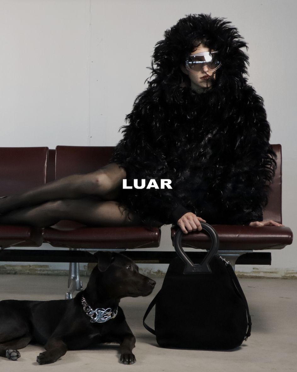 Person wearing a large black furry coat and sunglasses sitting on a red bench, with a black dog lying underneath, and a black handbag on the floor.