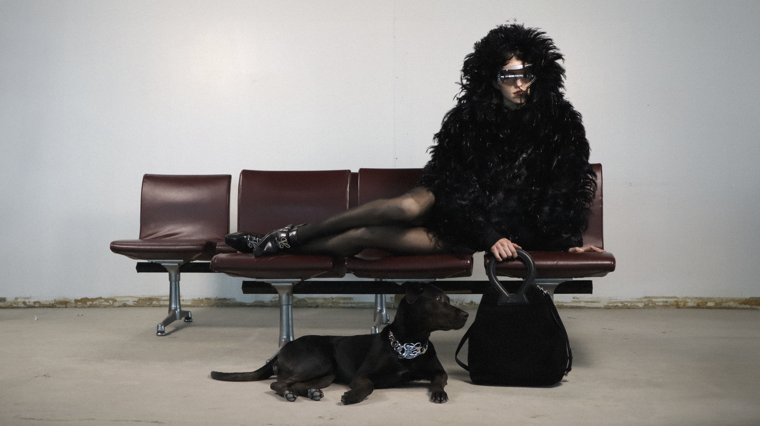 A woman with dark hair and sunglasses sitting on a row of red airport-style seats, wearing a large black faux fur coat, black tights, and black shoes with buckles. She has a black bag beside her, and a black dog with a chain collar is lying on the floor in front of her.