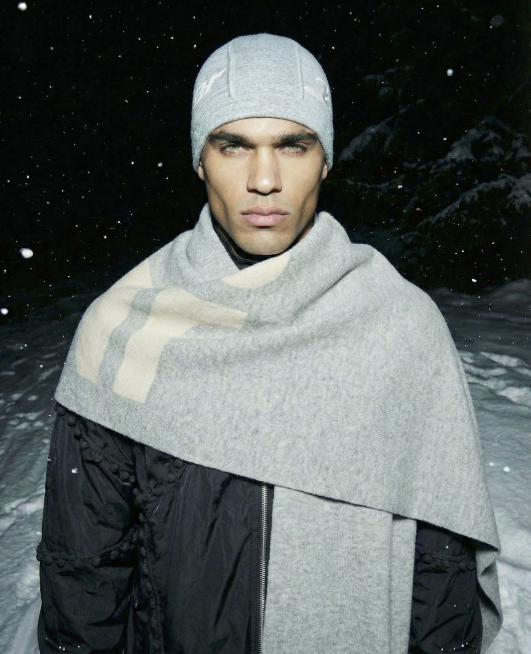 A young man with light skin, wearing a gray beanie, scarf, and black jacket, standing outdoors in a snowy, dark environment with snowflakes falling around him.