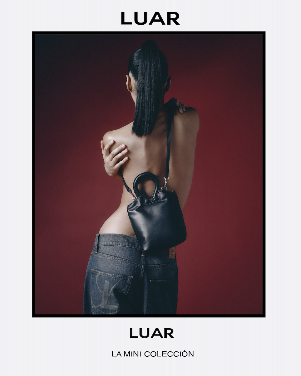 A woman with long dark hair in a high ponytail, topless and facing away, with one arm crossed over her chest and the other hand resting on her shoulder, wearing dark jeans and carrying a small black handbag against a red background.