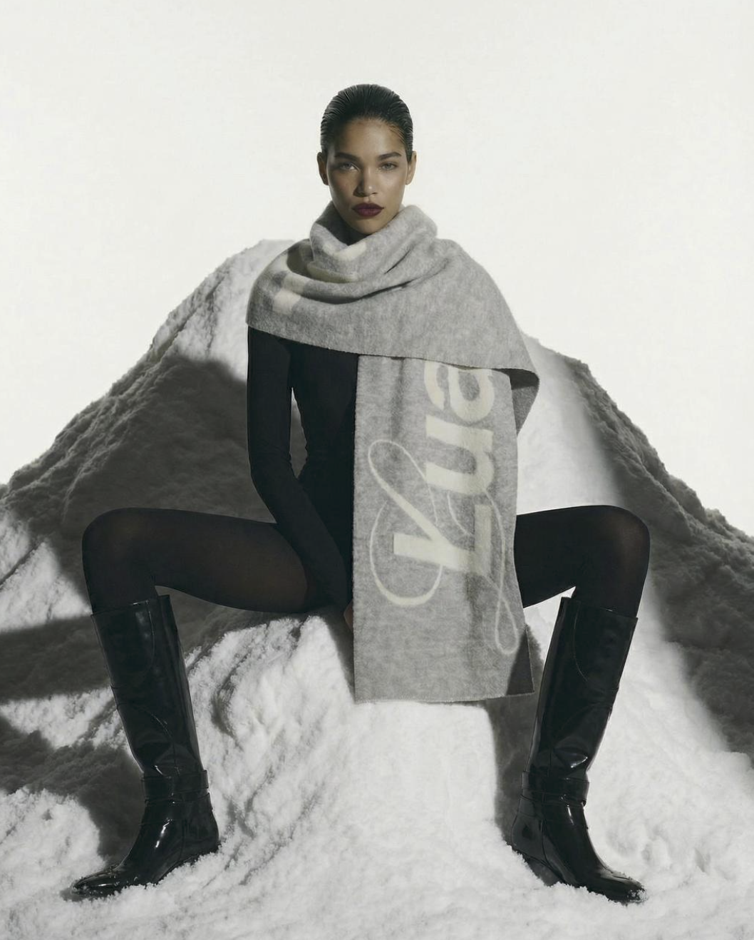A woman sitting on snow with legs apart, wearing black tights, black boots, a black long-sleeve top, and a large grey scarf with white lettering, against a plain light background.