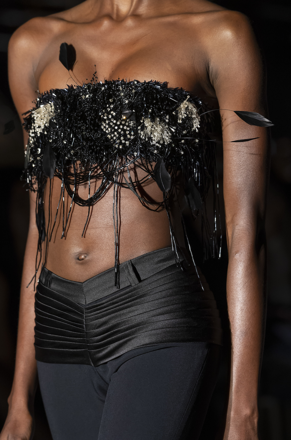 Close-up of a model wearing a strapless black top decorated with black feathers, beads, and embellishments, paired with high-waisted black pants with horizontal pleats.
