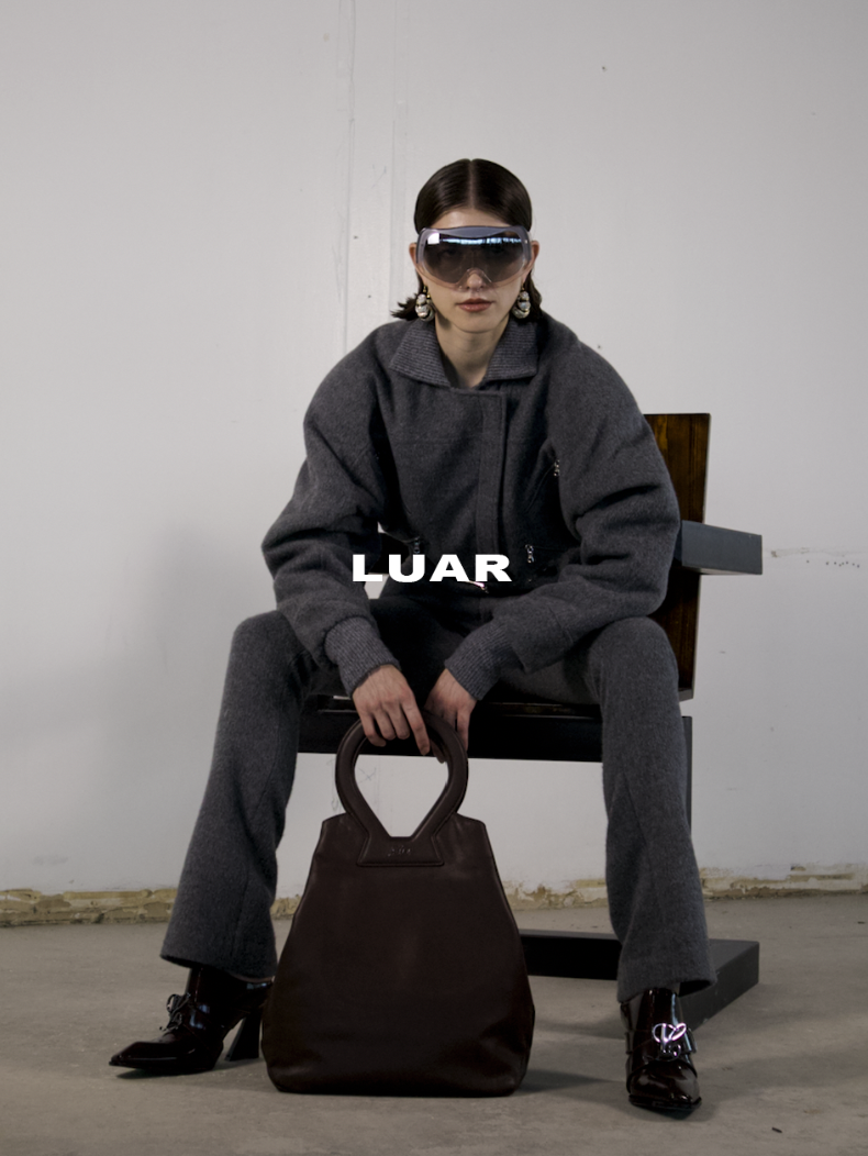 A woman sitting on a black chair wearing a dark gray oversized coat and matching gray pants, black high-heeled shoes, large sunglasses, and earrings, holding a large brown handbag in her lap, in an industrial setting with a plain white wall background.