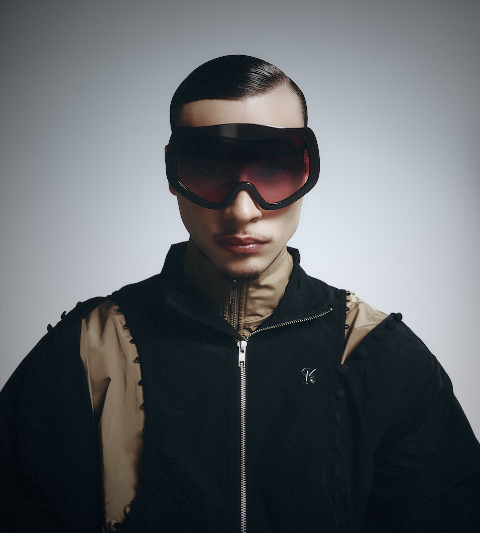 A person in futuristic black and beige clothing wearing large black and red goggles, with a sleek hair style against a plain background.