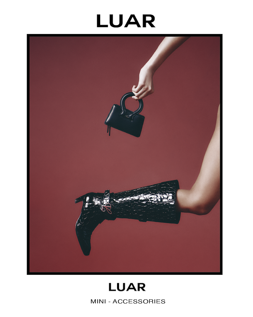 Fashion advertisement featuring a hand holding a small black purse and a leg wearing a shiny black high-heeled boot against a red background. Text reads "LUAR" at the top and bottom, with "MINI - ACCESSORIES" at the bottom.