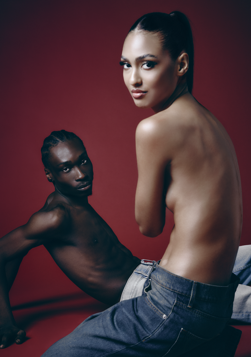 Two topless models, one with dark skin and braided hair reclining against a red background, and the other with light skin and long dark hair tied back, sitting with their backs slightly turned.