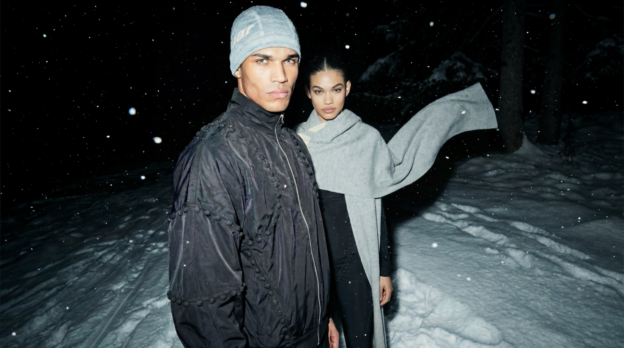 Two models standing in a snowy forest at night, dressed in winter clothing, with snow falling around them.