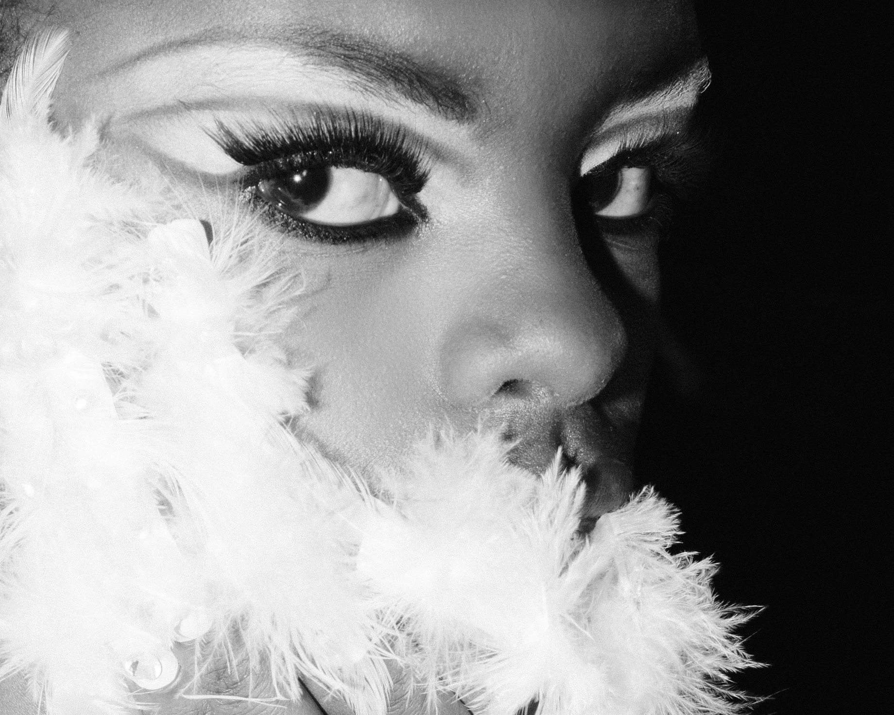 A close-up black-and-white photograph of a woman’s face, featuring her eye with dramatic makeup and a feathered accessory partially covering her face.