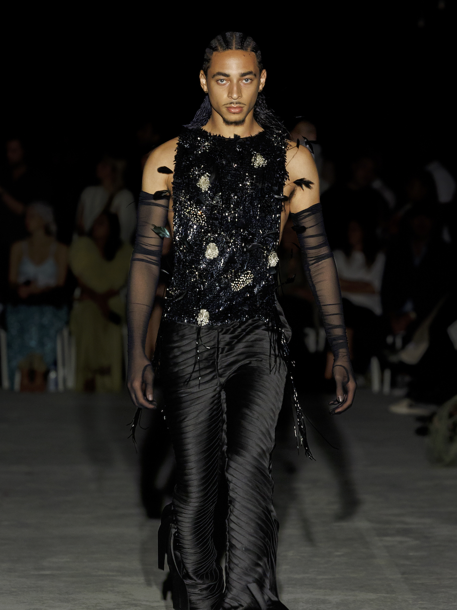 Model walking down runway in black, textured, and embellished outfit with sheer gloves at fashion show.