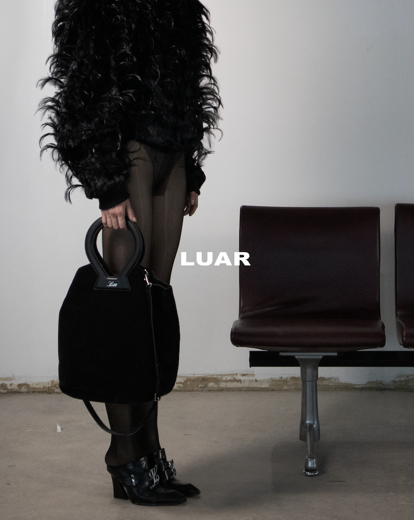 Person with long curly black hair, wearing brown pants, black top, and black boots, holding a black handbag, standing in an empty room with two dark red chairs.