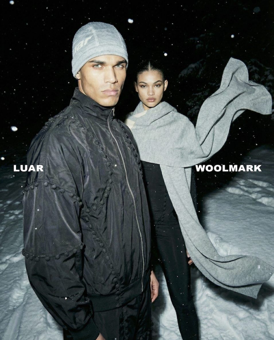 Two models standing in snow at night, wearing winter clothing. The male model on the left wears a gray beanie and black jacket. The female model on the right wears a large gray scarf and a gray coat.