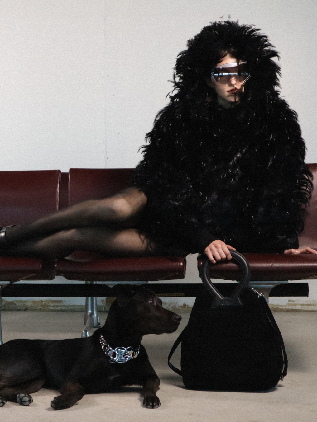 A woman dressed in a black fuzzy coat and sunglasses seated on a red bench, with a small black dog lying underneath her. The woman is wearing black tights and has short dark hair. A black handbag is on the floor beside the dog, and the background is plain and simple.