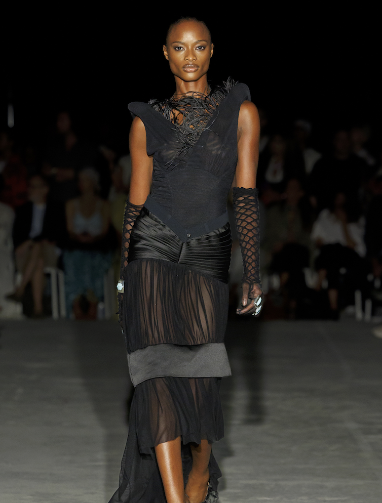 A Black female model walking on a runway in a black sheer dress with layered ruffles and textured details, with an audience in the background.