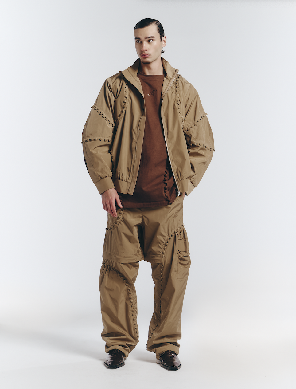 Young man wearing a tan track suit with decorative stitching, brown T-shirt, and black shoes, standing against a white backdrop.