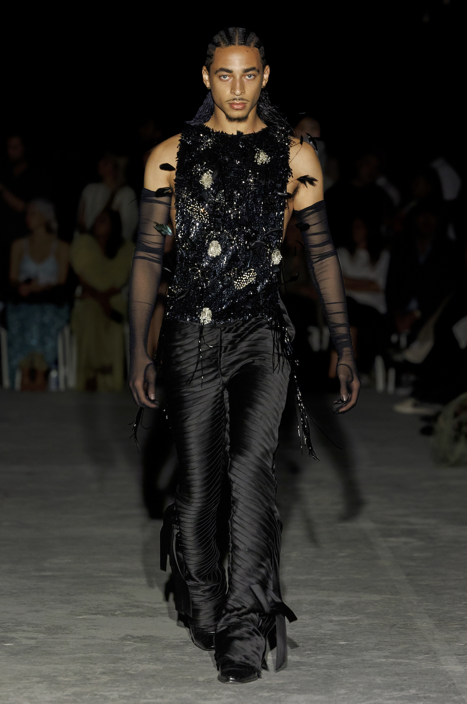 Model walking down runway in black, textured, and embellished outfit with sheer gloves at fashion show.