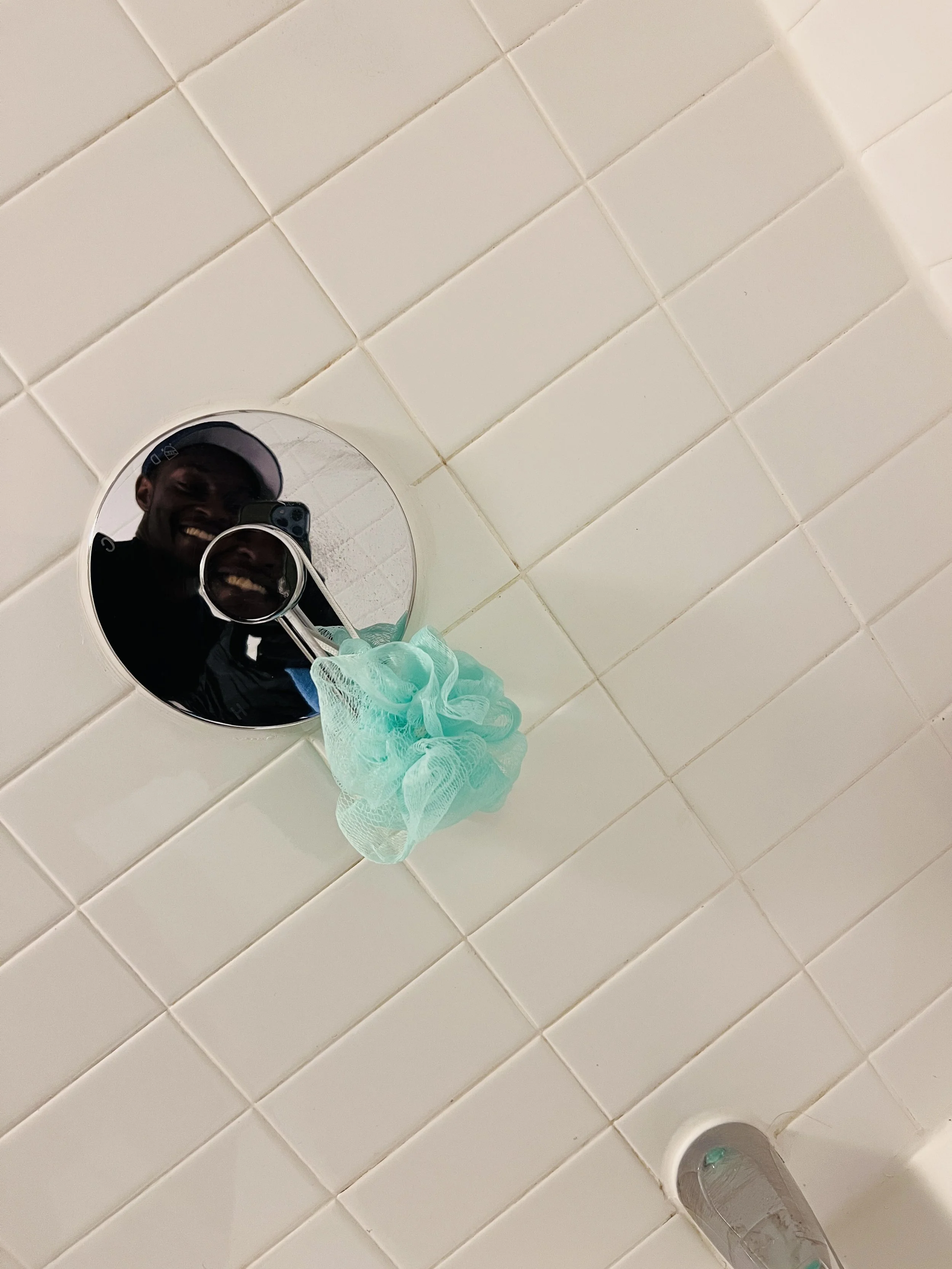 Reflection of a person smiling while taking a selfie in a bathroom mirror, with white tiled wall and a blue bath sponge hanging from the mirror.