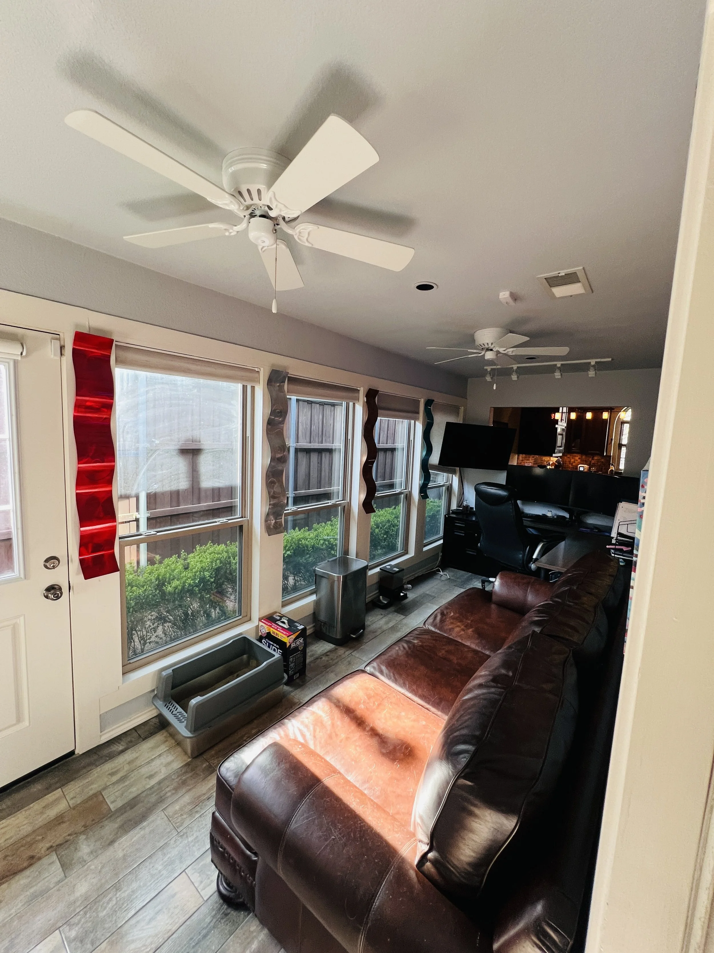 Living room with a brown leather sofa, a window with red and gray twisted paper decorations, a ceiling fan, and a flat-screen TV above a desk with an office chair.