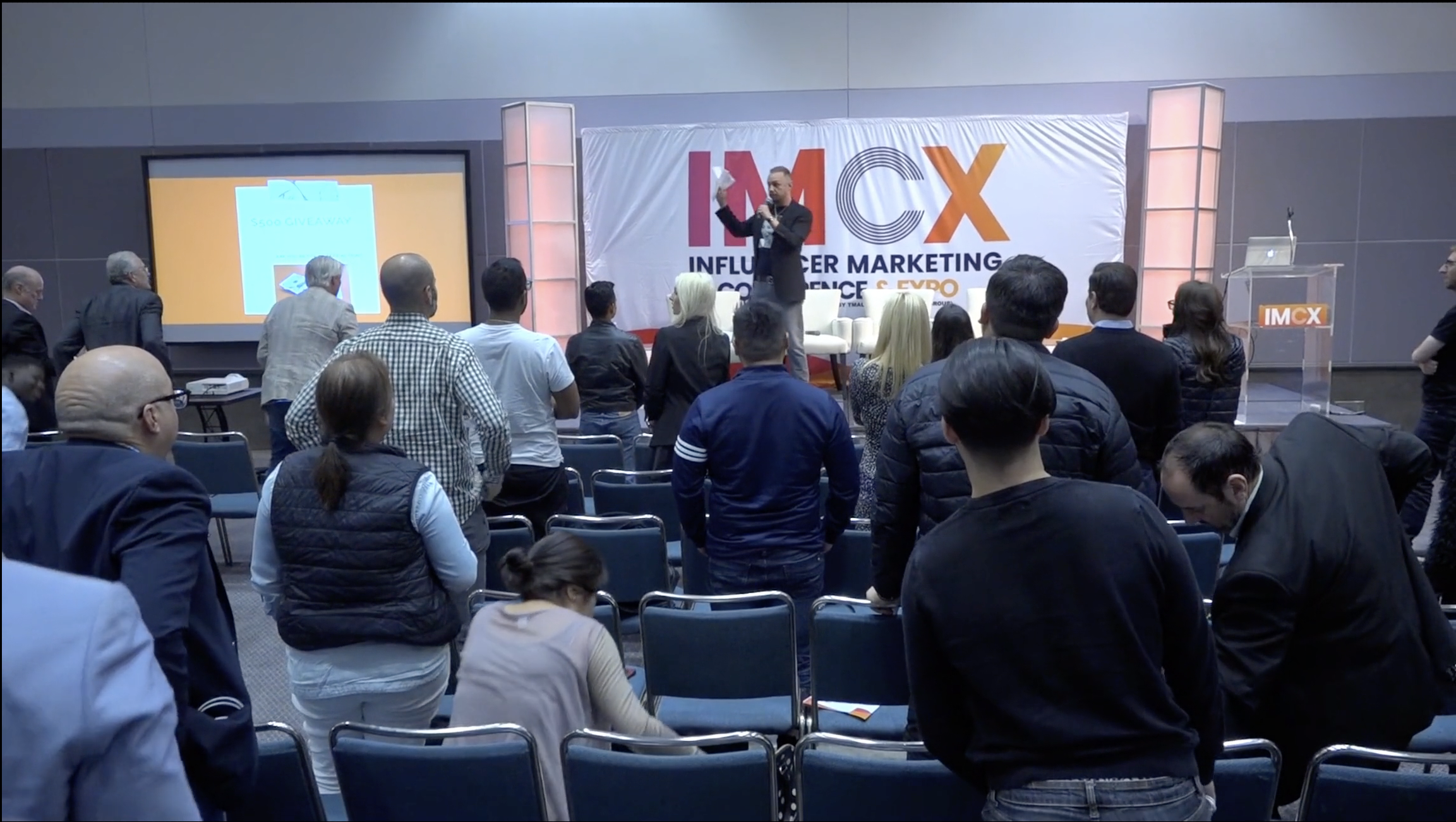 People gathered at a conference in a large room, listening to a presenter on stage with a backdrop reading 'IMCX Influencer Marketing Conference.' A screen displays a slide about a giveaway.