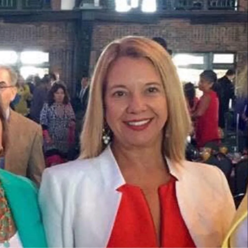 A woman with blonde hair wearing a white blazer and red top, smiling, at an indoor event with people in the background.