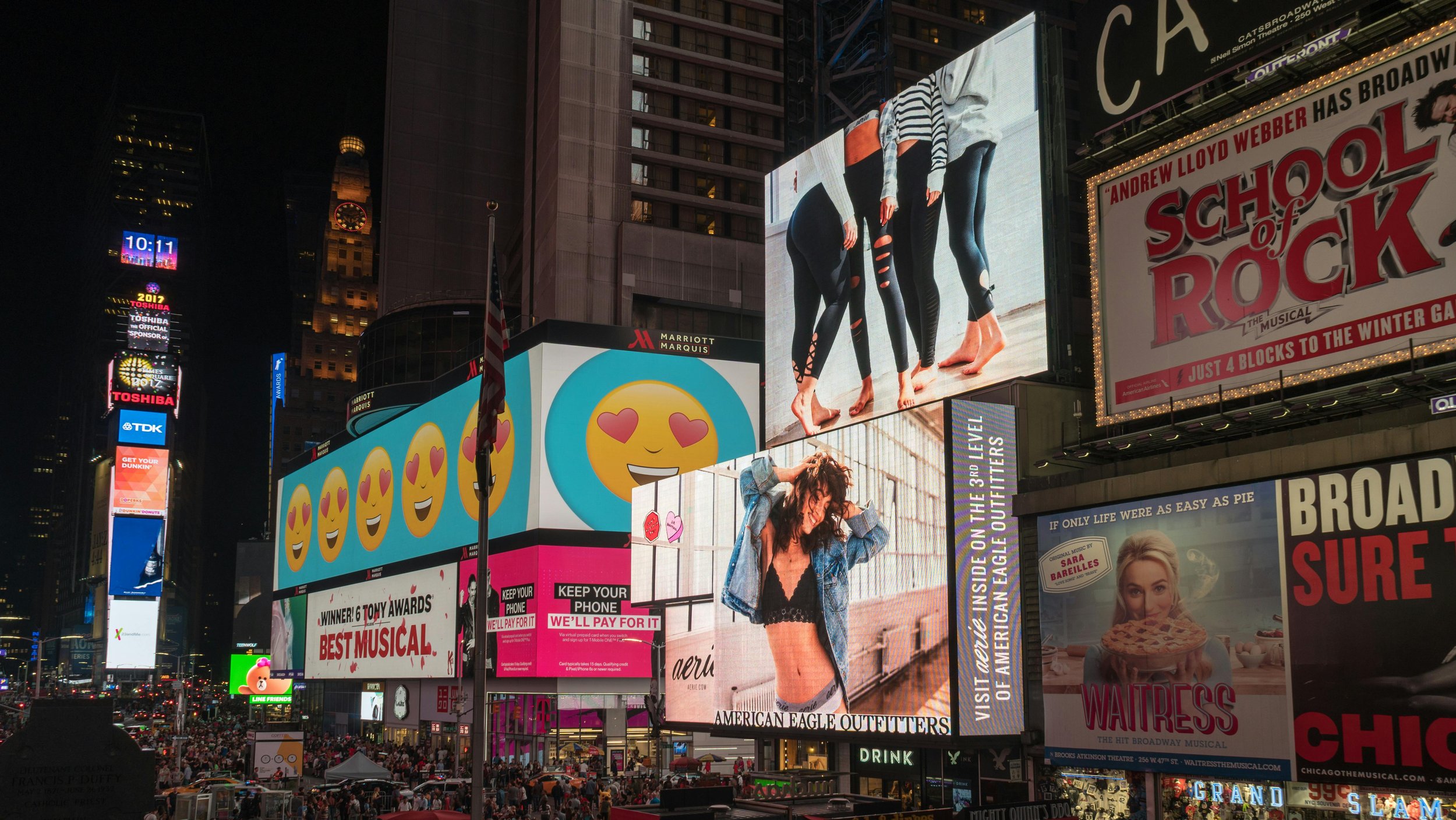 Brightly lit billboards and digital screens in Times Square at night, with advertisements for various movies, brands, and products, and a crowd of people below.