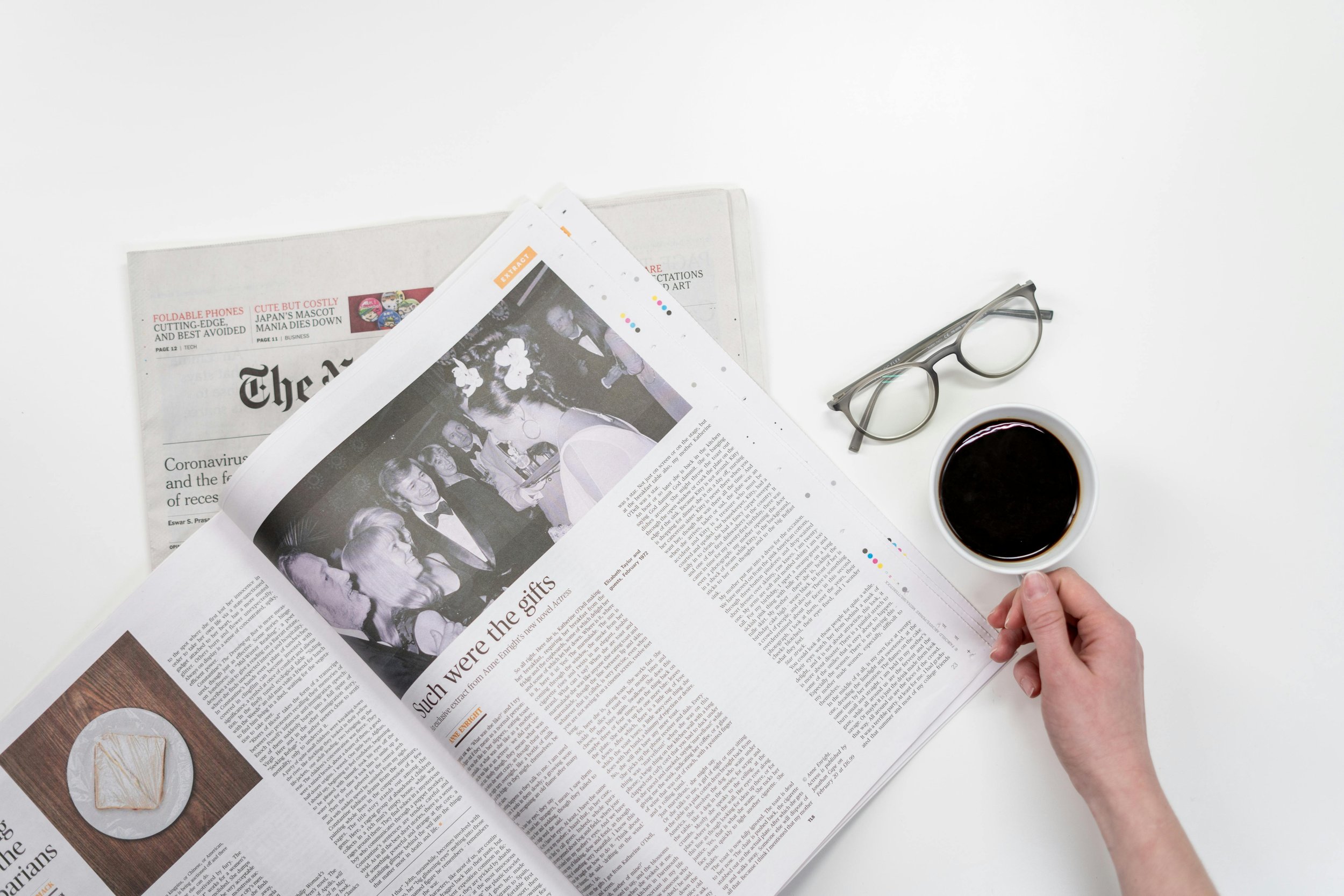 Top view of a person's hand holding a cup of coffee on a white desk, with newspaper, a pair of glasses, and additional newspapers nearby.