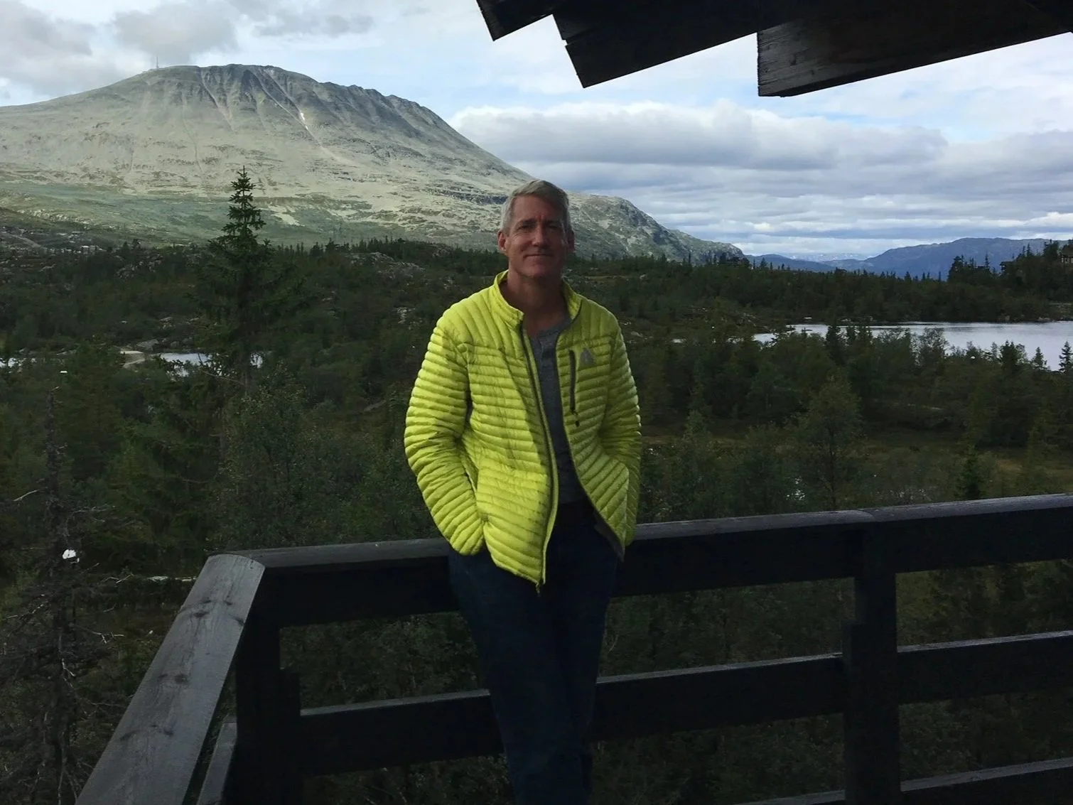 A man in a neon yellow jacket standing on a wooden railing overlooking a landscape with trees, water, mountains, and cloudy sky.