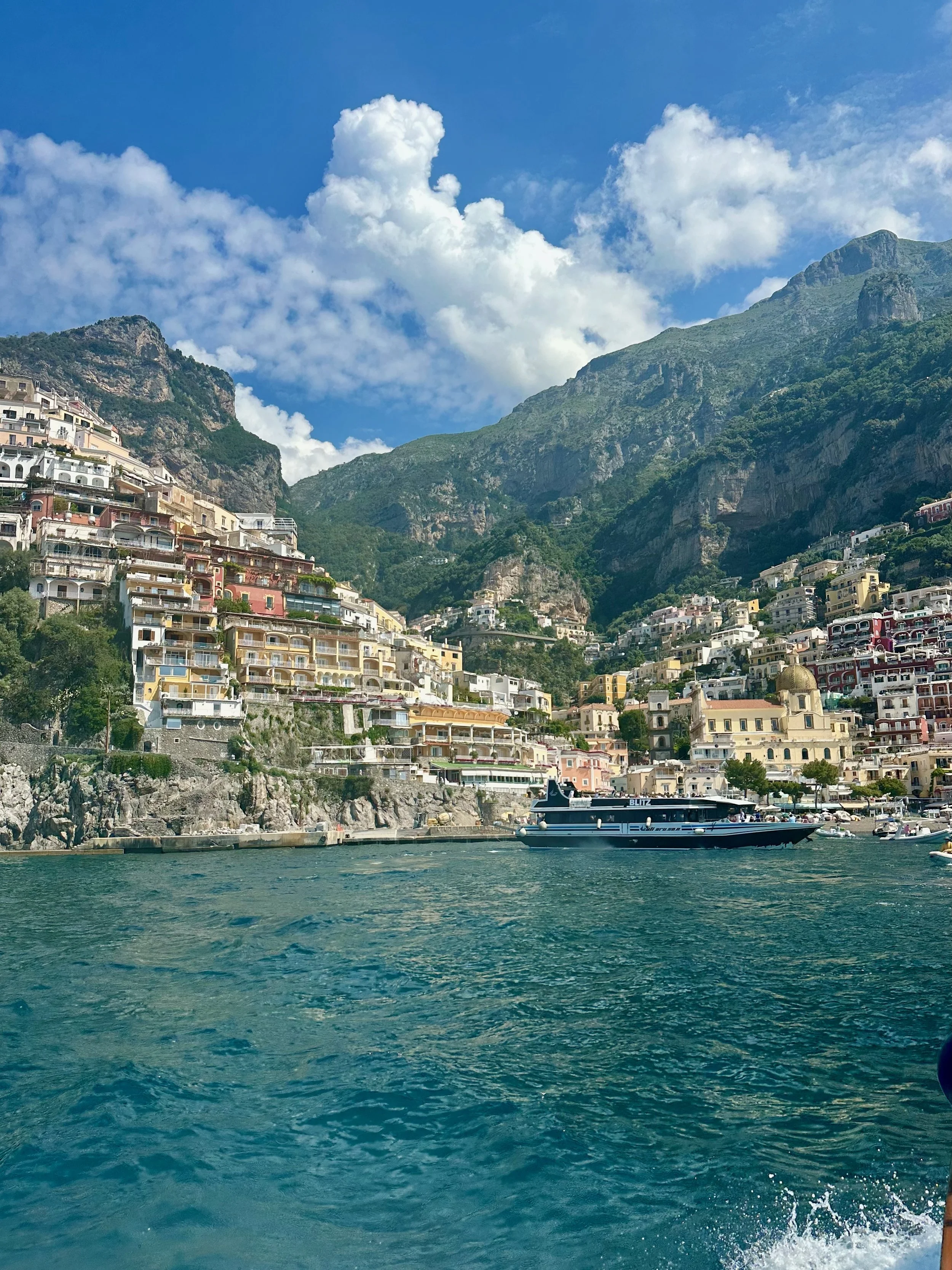 Scenic view of a coastal city with colorful buildings on steep hillsides, mountains in the background, and a body of water with boats in the foreground under a partly cloudy sky.