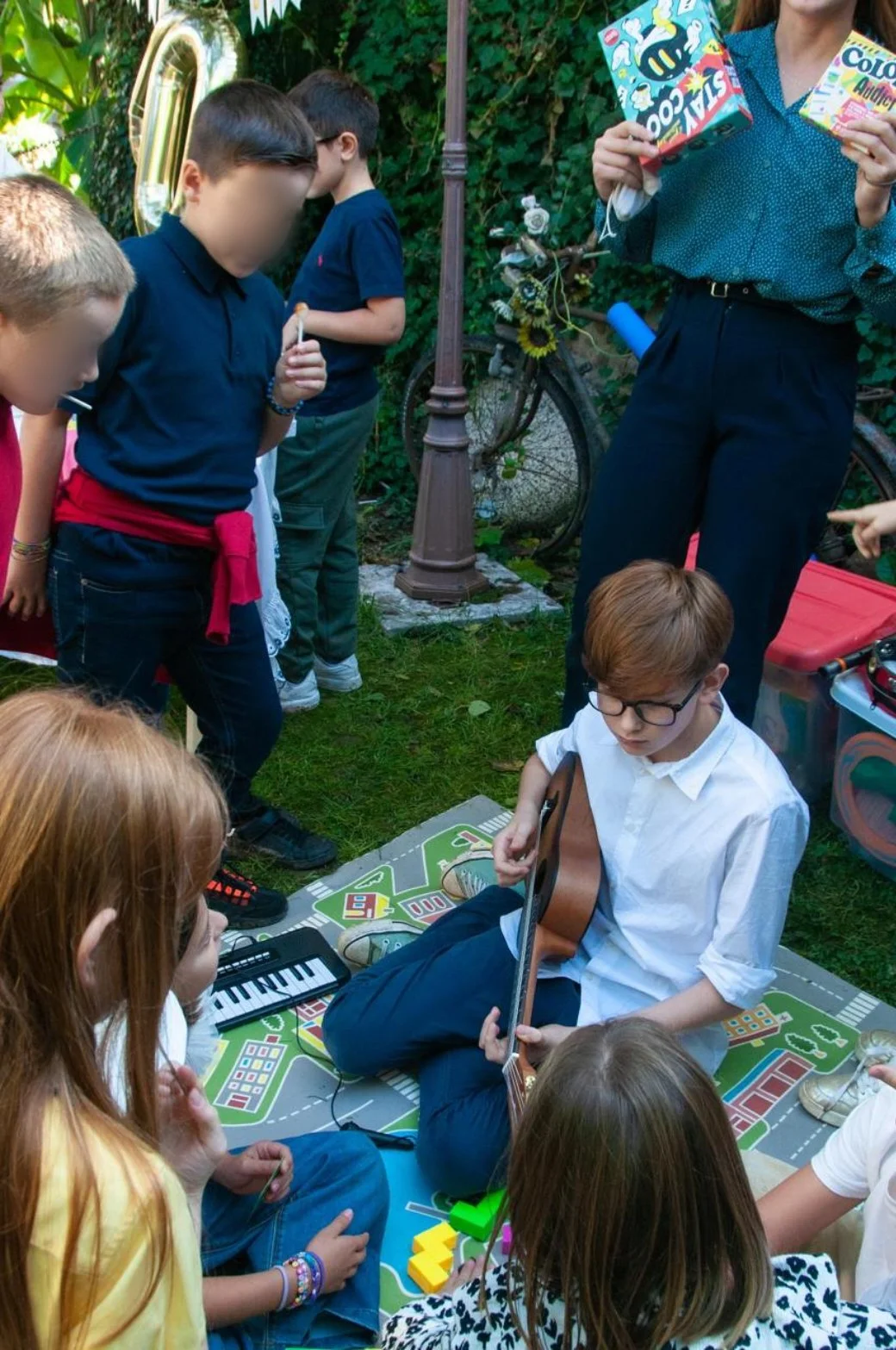 Gruppo di bambini e un adulto in un giardino, alcuni che ascoltano e altri che suonano la chitarra, in una scena di ritrovo e musica all'aperto.