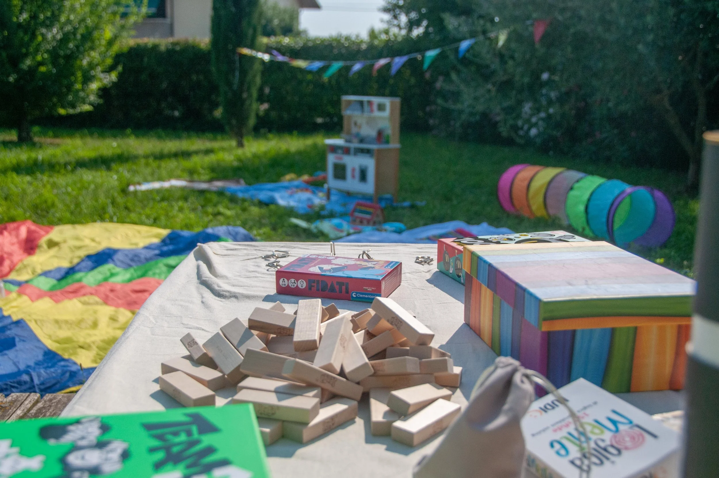 Gioco di società composto da pezzi di legno, una scatola colorata e altre scatole con giochi da tavolo, in un'area esterna con erba e alberi.