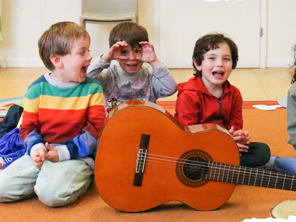 Tre bambini seduti sul tappeto con una chitarra classica davanti, che fanno facce divertenti e sorridono.