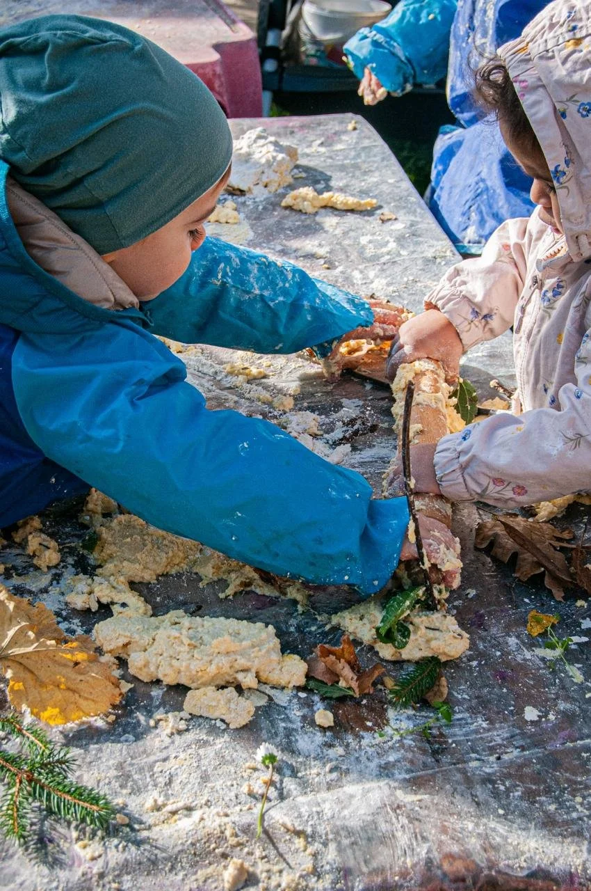Due bambini vestiti con abiti invernali che impastano qualcosa sulla tavola all'aperto. La scena è autunnale, con foglie secche e qualche pianta sul tavolo.