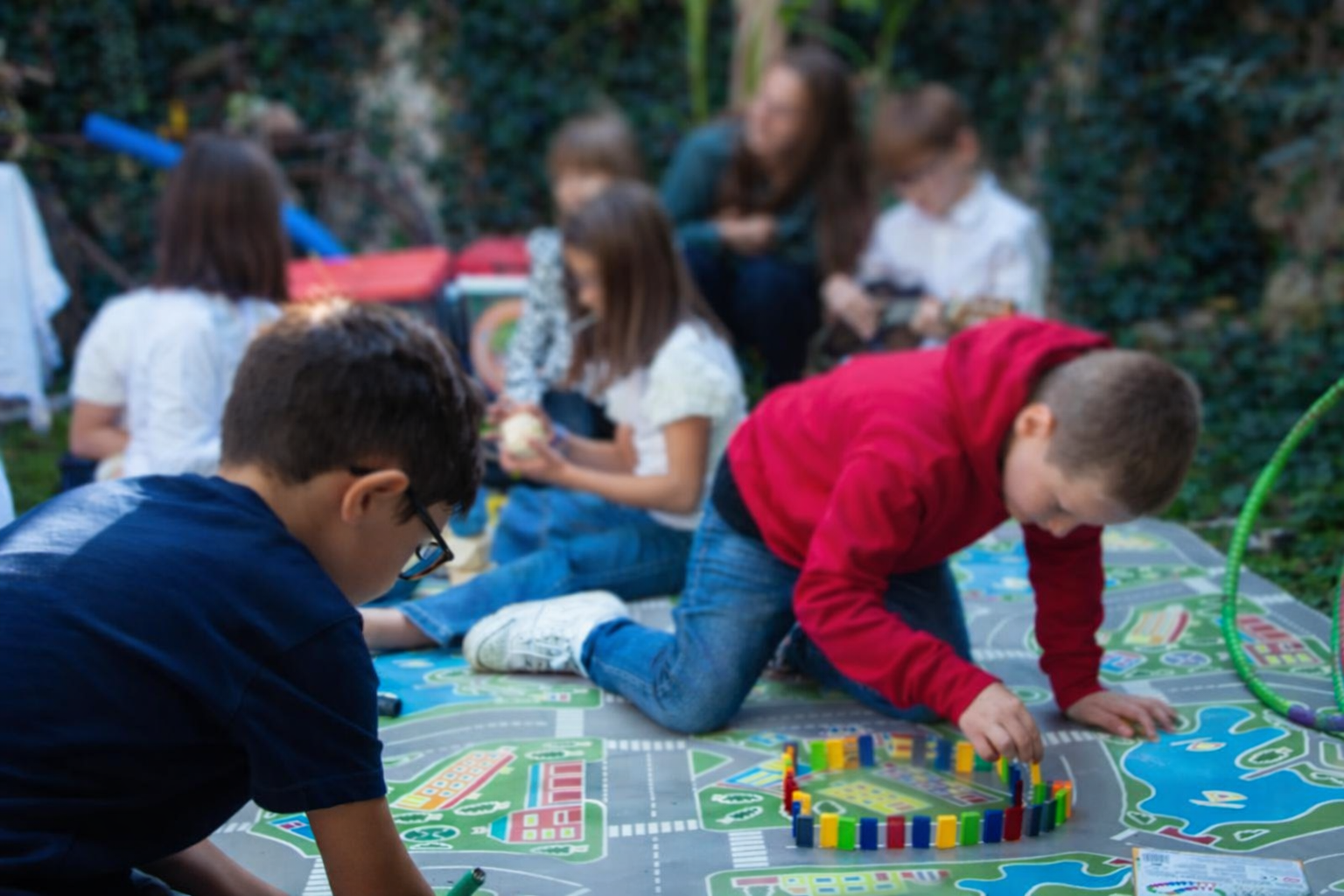 Gruppo di bambini che giocano e si divertono all'aperto su un grande tappeto con un percorso e un gioco di costruzioni, con alcune bambine svolgendo attività di lettura e un adulto che supervisiona.