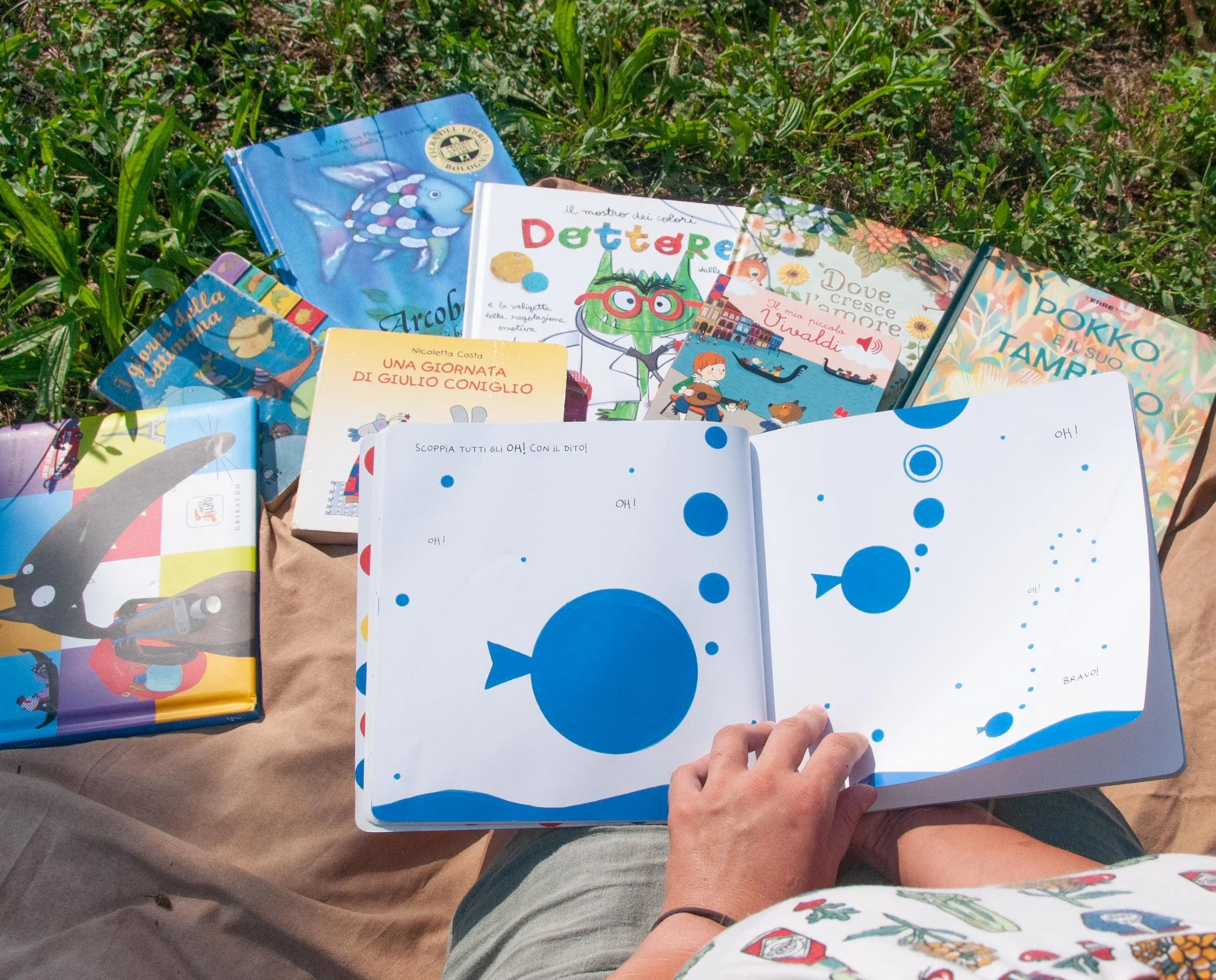 Una persona tiene un libro en las manos, mientras otros libros están dispuestos sobre una mesa en un entorno al aire libre con césped y tierra.
