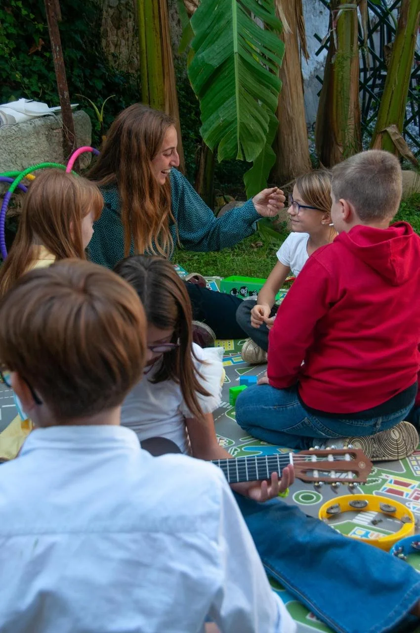Un gruppo di bambini seduti su un tappeto con una donna che suona un fiocco, in un ambiente all'aperto con piante verdi.