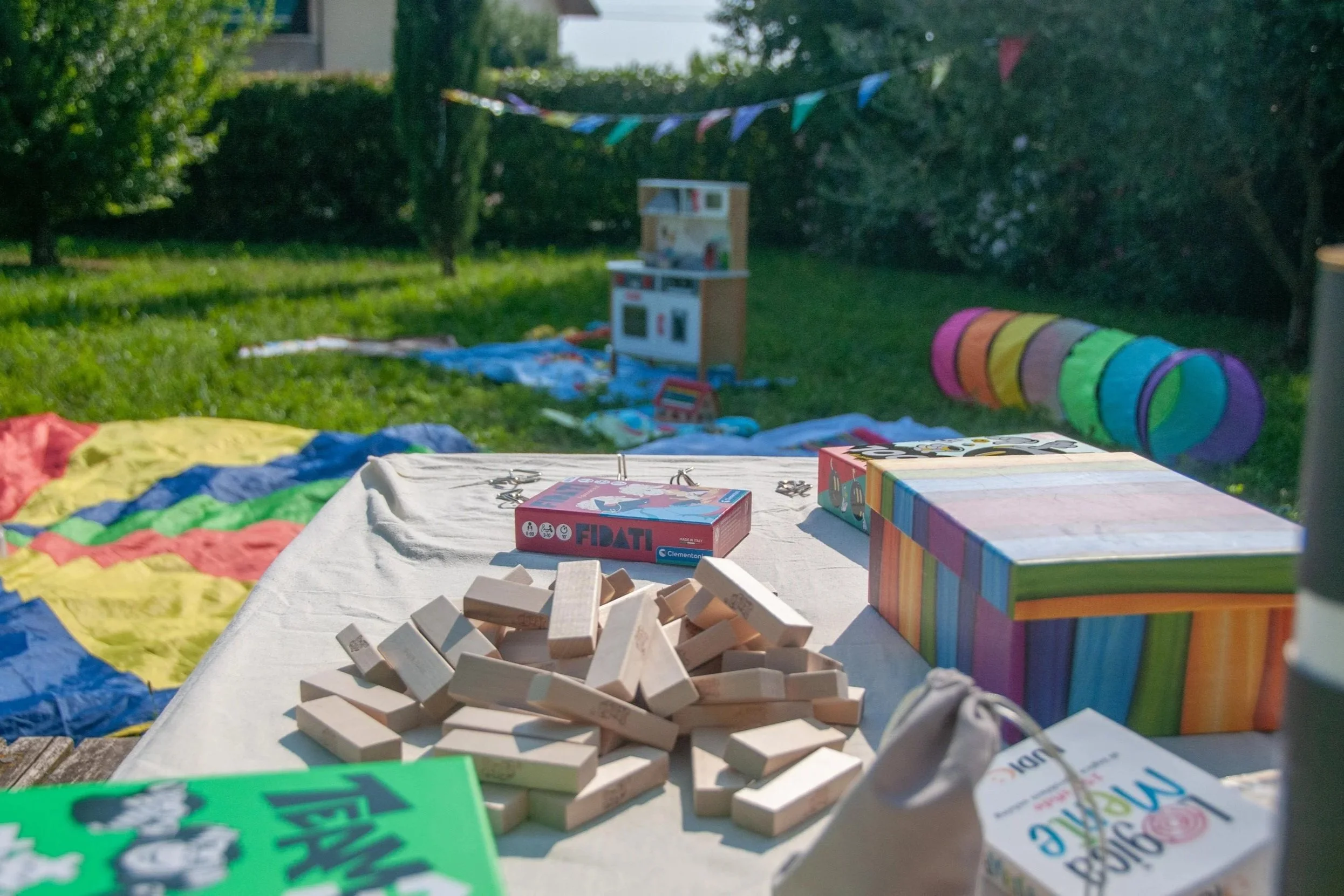 Gioco di costruzioni in legno e vari giochi all'aperto su un tavolo in giardino, con decorazioni di bandiere colorate sospese tra gli alberi.