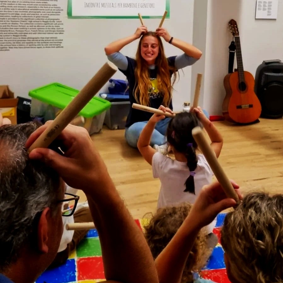 Una ragazza sorridente con orecchie fatte con bacchette di legno insegna a bambini in un laboratorio musicale, con chitarra e altri strumenti sullo sfondo.