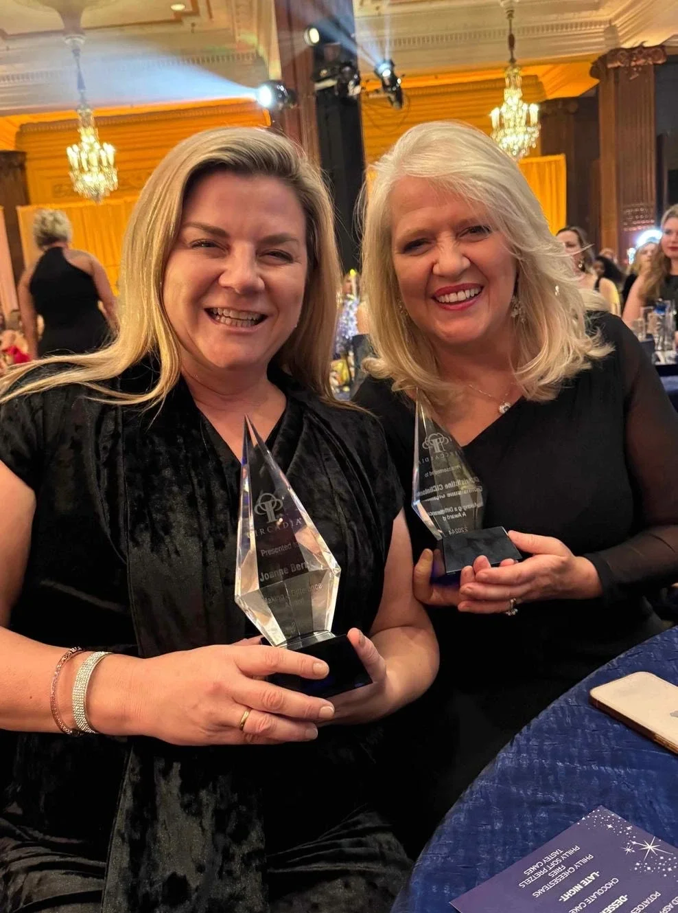 Two women in black dresses smiling and holding crystal awards at an elegant event with chandeliers and golden curtains in the background.