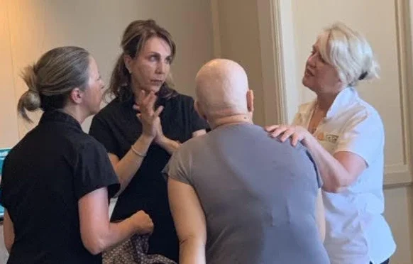 Four people, two women and two men, engaged in a serious conversation in a room with beige walls. One woman has blonde hair and glasses, another woman has brown hair, and the two men are bald, one with a gray shirt and the other with a dark shirt. The women are gesturing with their hands while speaking.