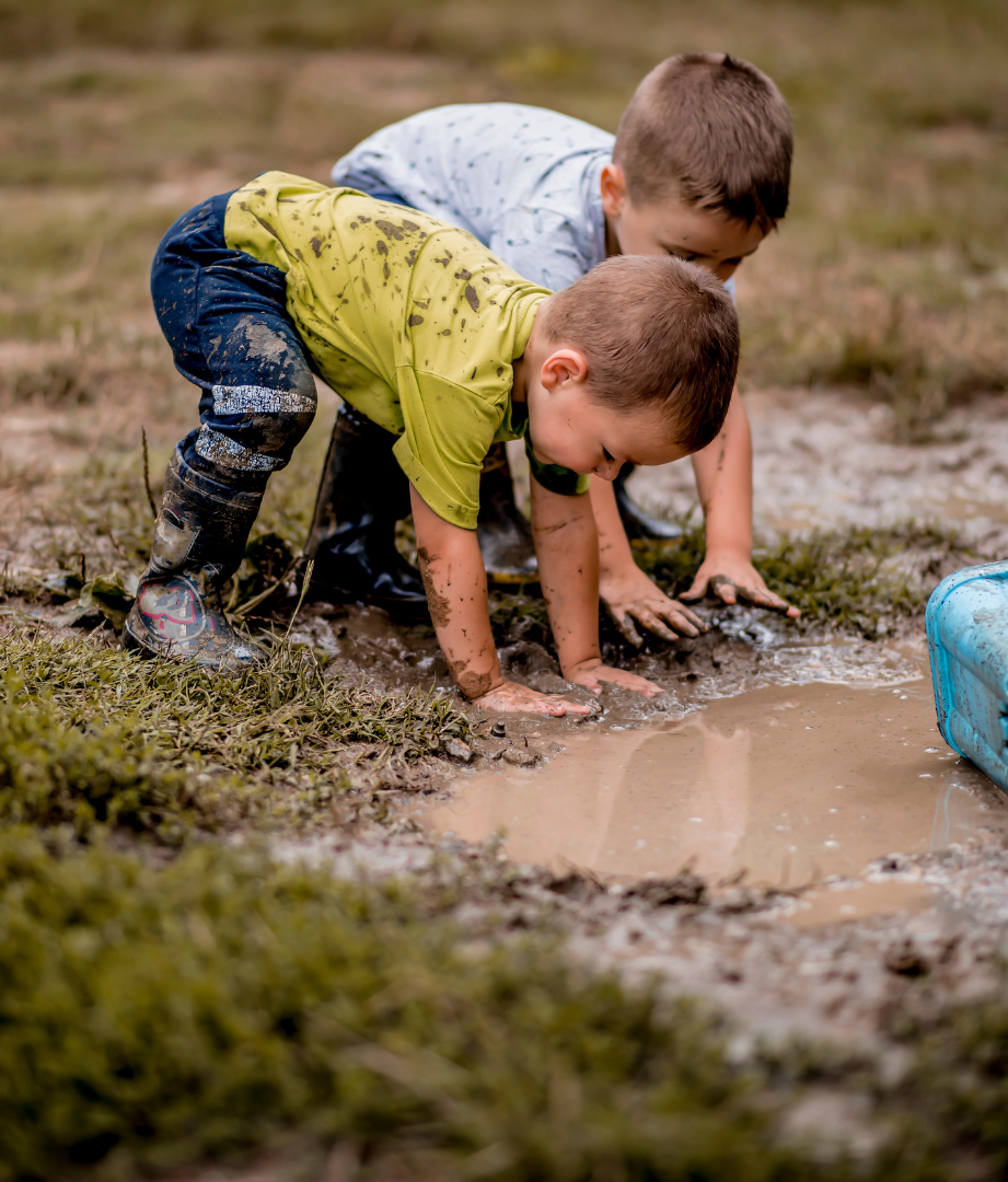 Two young boys playing in a muddy puddle outdoors, bending over and touching the muddy water with their hands and knees.