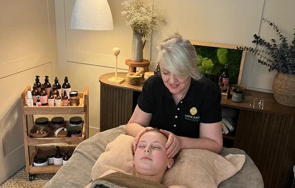 A woman giving a facial treatment to a young boy lying on a massage table in a cozy, softly lit room with shelves of skincare products and decorative plants.