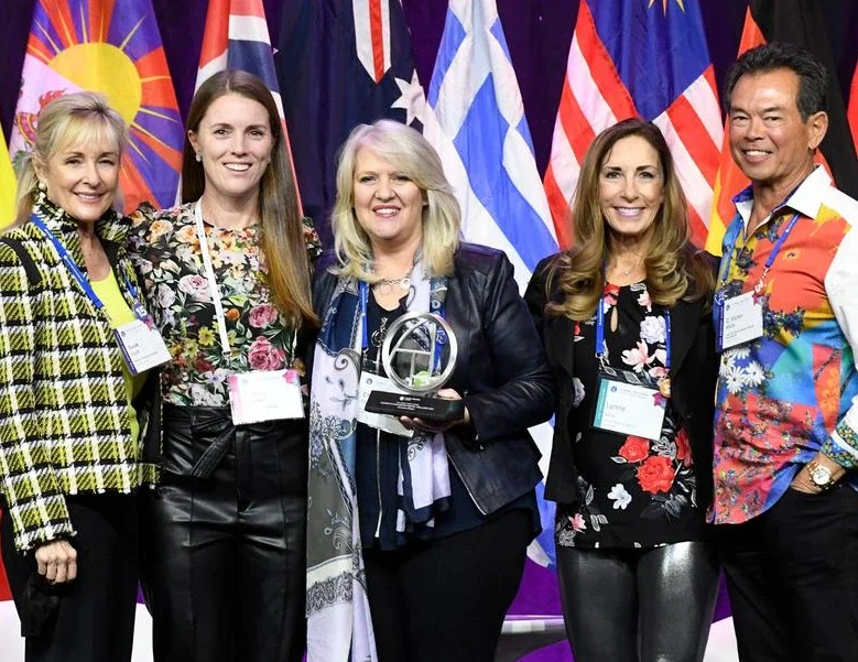Group of five women and one man at an awards event with flags in the background, one woman holding a glass award.
