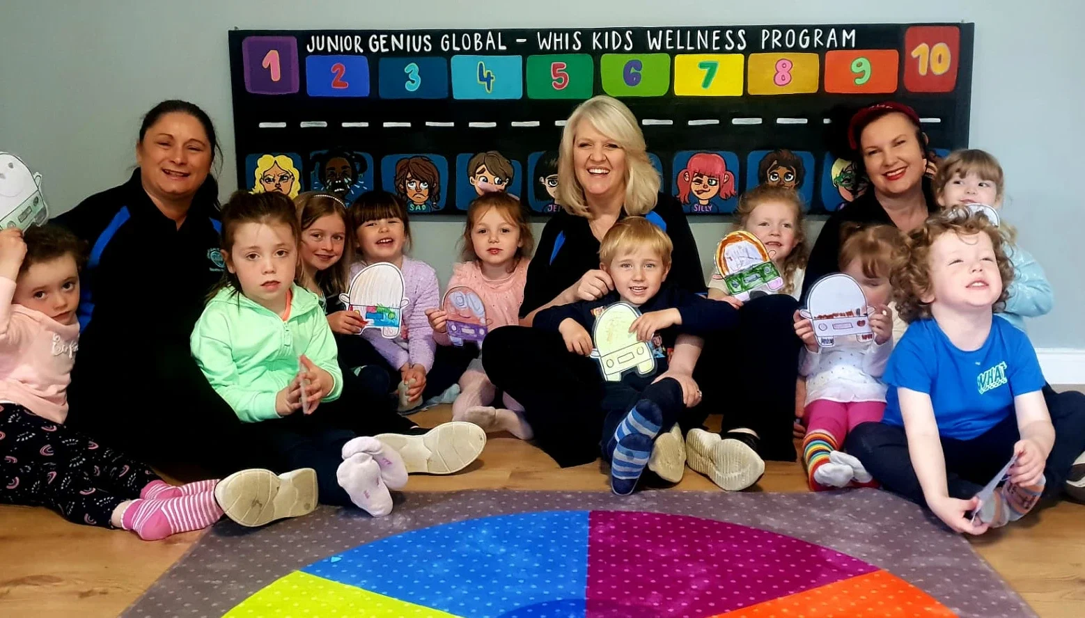 Group of young children and two adult women sitting on the floor in a classroom, holding paper craft cars, with a colorful learning chart and cartoon faces on the wall behind them.