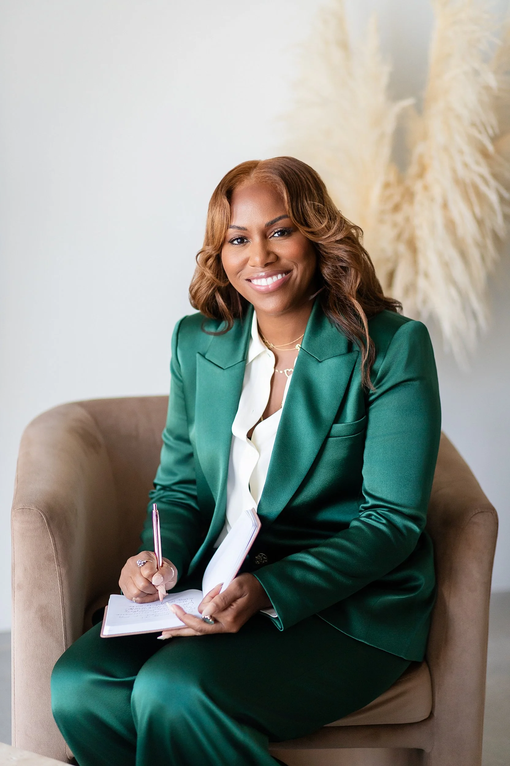 A woman with brown hair and a green blazer sitting on a beige chair, smiling, holding a notebook and pen, with neutral decor in the background.