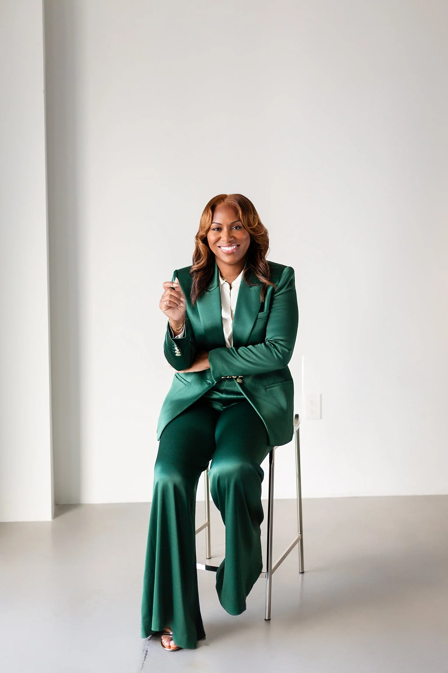 A woman wearing a green suit sitting on a stool with a bright smile, in front of a plain white background.